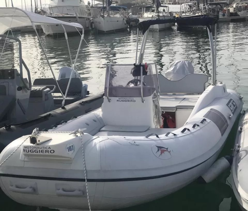 Un gommone bianco con consolle centrale, tientibene e roll-bar, ormeggiato tra le barche di un porto turistico